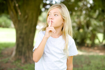 Fototapeta premium Caucasian little kid girl wearing whiteT-shirt standing outdoors silence gesture keeps index finger to lips makes hush sign. Asks not to share secret