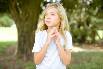 Caucasian little kid girl wearing whiteT-shirt standing outdoors steepls fingers and looks mysterious aside has great evil plan in mind