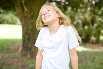 Fototapeta premium Positive Caucasian little kid girl wearing whiteT-shirt standing outdoors with overjoyed expression closes eyes and laughs shows white perfect teeth