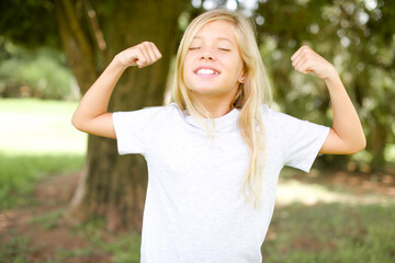 Strong powerful Caucasian little kid girl wearing whiteT-shirt standing outdoors toothy smile, raises arms and shows biceps. Look at my muscles!