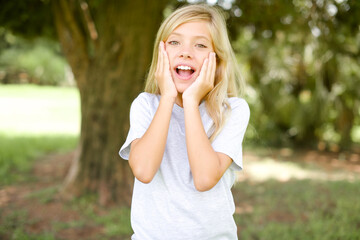 Caucasian little kid girl wearing whiteT-shirt standing outdoors Pleasant looking cheerful, Happy reaction