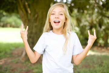 Caucasian little kid girl wearing whiteT-shirt standing outdoors makes rock n roll sign looks self...