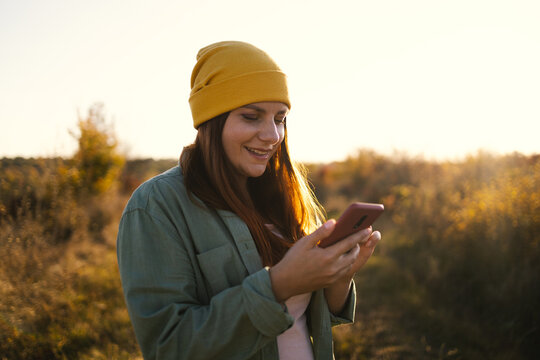 Stylish 30s caucasian female in a bright yellow knitted hat using phone in the field. Beautiful autumn nature background - Powered by Adobe
