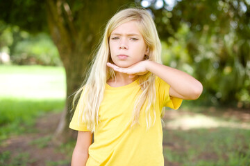 beautiful Caucasian little kid girl wearing yellow T-shirt standing outdoors cutting throat with...
