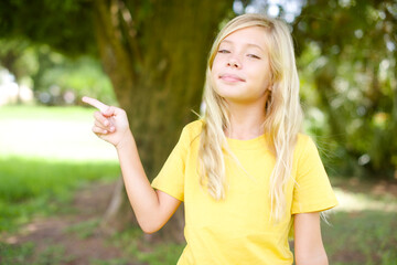Positive beautiful Caucasian little kid girl wearing yellow T-shirt standing outdoors with satisfied expression indicates at upper right corner shows good offer suggests to click on link