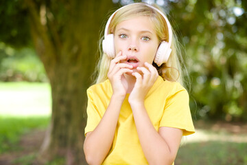 Shocked beautiful Caucasian little kid girl wearing yellow T-shirt standing outdoors stares fearful at camera keeps mouth widely opened wears wireless stereo headphones on ears