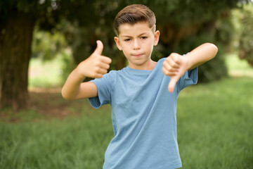 beautiful Caucasian little kid boy wearing blue T-shirt standing outdoors showing thumbs up and thumbs down, difficult choose concept - obrazy, fototapety, plakaty
