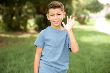 beautiful Caucasian little kid boy wearing blue T-shirt standing outdoors Waiving saying hello...