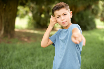 beautiful Caucasian little kid boy wearing blue T-shirt standing outdoors smiling cheerfully and pointing to camera while making a call you later gesture, talking on phone