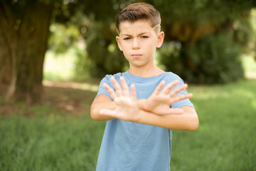 beautiful Caucasian little kid boy wearing blue T-shirt standing outdoors has rejection expression...