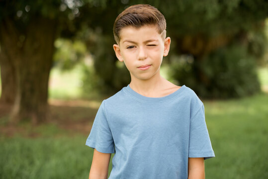 Coquettish beautiful Caucasian little kid boy wearing yellow T-shirt standing outdoors smiling happily, blinking at camera in a playful manner, flirting with you.