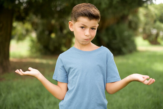 Clueless beautiful Caucasian little kid boy wearing yellow T-shirt standing outdoors shrugs shoulders with hesitation, faces doubtful situation, spreads palms, Hard decision