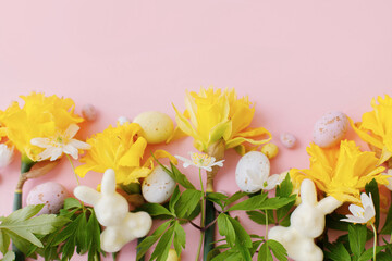Happy Easter! Colorful Easter chocolate eggs, bunnies and daffodils flowers border on pink background. Stylish Easter flat lay. Greeting card or banner template