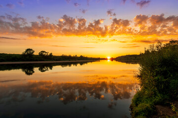 Naklejka na ściany i meble Amazing view at scenic landscape on a beautiful river and colorful sunset with reflection on water surface and glow on a background, spring season landscape