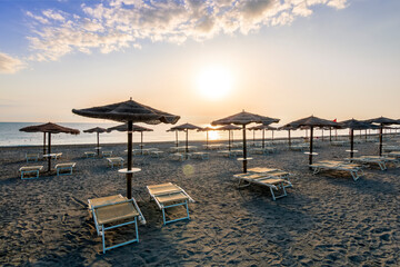 Fototapeta premium beautiful landscape of empty sunrise or sunset beach with chaise loungues and nice umbrellas with blue sea, sun glow and amazing cloudy sky on thr background