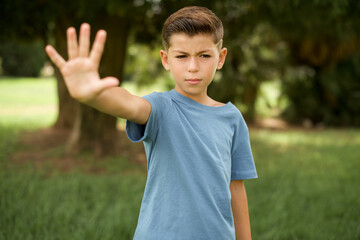 beautiful Caucasian little kid boy wearing blue T-shirt standing outdoors doing stop sing with palm of the hand. Warning expression with negative and serious gesture on the face.