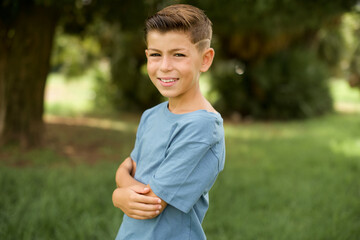 beautiful Caucasian little kid boy wearing blue T-shirt standing outdoors  happy face smiling with crossed arms looking at the camera. Positive person.