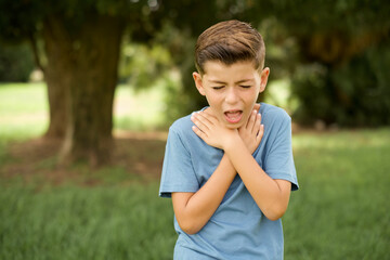 beautiful Caucasian little kid boy wearing blue T-shirt standing outdoors shouting suffocate because painful strangle. Health problem. Asphyxiate and suicide concept. - obrazy, fototapety, plakaty