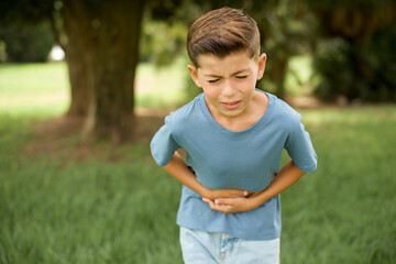 Caucasian little kid boy wearing blue T-shirt standing outdoor with hand on stomach because nausea,...
