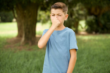 Caucasian little kid boy wearing blue T-shirt standing outdoor smelling something stinky and disgusting, intolerable smell, holding breath with fingers on nose. Bad smell