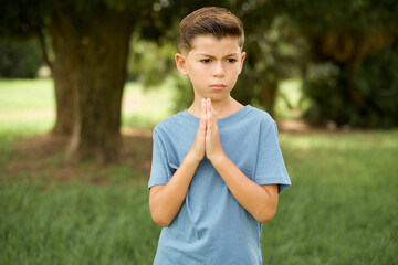 Caucasian little kid boy wearing blue T-shirt standing outdoor begging and praying with hands together with hope expression on face very emotional and worried. Please God