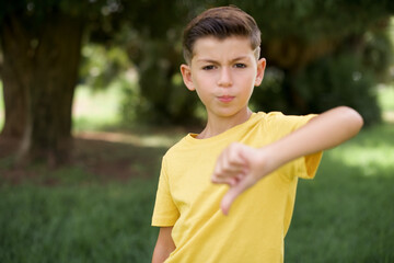 Caucasian little kid boy wearing yellow T-shirt standing outdoors looking unhappy and angry showing rejection and negative with thumbs down gesture. Bad expression.