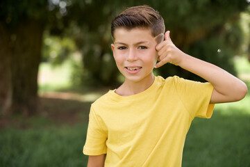 Caucasian little kid boy wearing yellow T-shirt standing outdoors makes phone gesture, says call me back again, has glad expression.