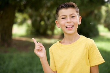 Caucasian little kid boy wearing yellow T-shirt standing outdoors laughs happily points away on blank space demonstrates shopping discount offer, excited by good news or unexpected sale.