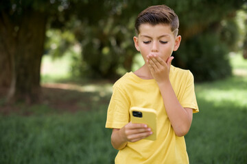 Caucasian little kid boy wearing yellow T-shirt standing outdoors being deeply surprised, stares at smartphone display, reads shocking news on website, Omg, its horrible!