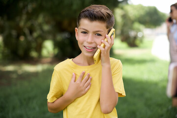 Fototapeta premium Smiling Caucasian little kid boy wearing yellow T-shirt standing outdoors talks via cellphone, enjoys pleasant great conversation. People, technology, communication concept
