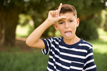 Funny beautiful Caucasian little kid boy wearing stripped T-shirt standing outdoors makes loser...
