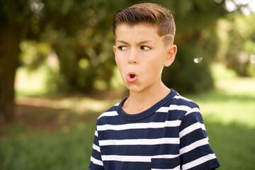 Shocked beautiful Caucasian little kid boy wearing stripped T-shirt standing outdoors look empty space with open mouth screaming: Oh My God! I can't believe this.