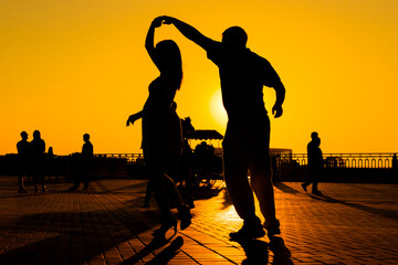 Woman and man couple silhouettes dancing against warm sunset orange sky on quay at evening. Group dance, romantic, love, summer outdoor activity and vacation concept
