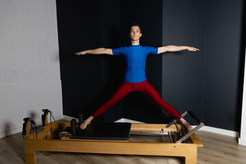 Sporty man doing exercises on pilates reformer training machine. Working out wearing sportswear, gym.