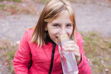 Young girl after an active walk in the park drinks clean water from a bottle and smiles, the girl is happy after jogging in the fresh air
