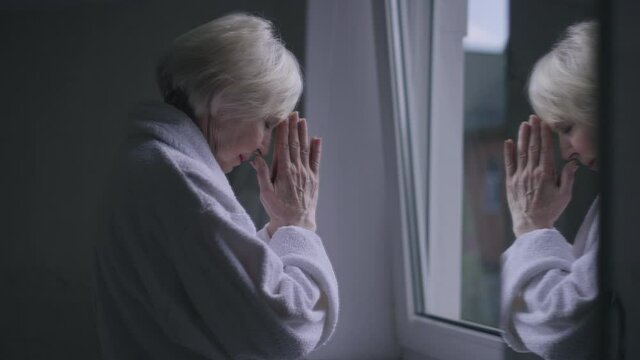 Side view reflection in mirror of sad senior Caucasian woman praying standing at window indoors. Upset depressed lonely retiree in bathrobe with hands together asking God for help and thanking
