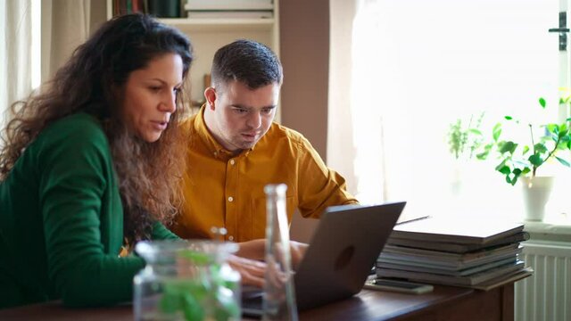 Young man with Down syndrome with his tutor studying and using laptop indoors at home, homeschooling.