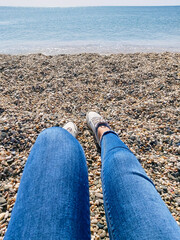 A woman is sitting on the seashore. Woman legs in white sneakers and jeans on grey pebble beach in summer day. Ocean clear water, small waves, good weather. Summer marine background.