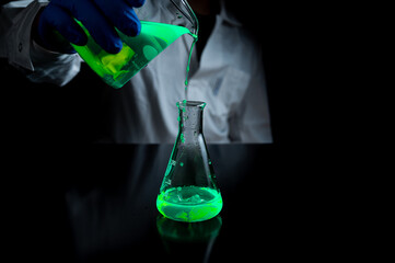 A woman scientist experimenting with a green fluorescent solution in a glass conical flask in dark...