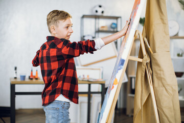 Inspired caucasian boy creating his masterpiece on easel using paints and hands. Little student enjoying art lesson at modern school.