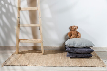 The teddy bear is leaning against the wall next to a small children's stool with three gray pillows on it. The stepladder is leaning against the wall