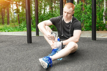 A young athlete is resting on the playground with a bottle of water and a phone. The athlete sitting on the map is looking at his smartphone.