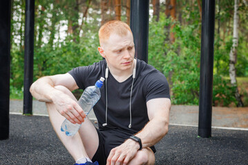 Young man with towel on his shoulders drinking water while sitting at street.