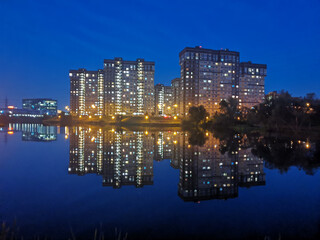 Multi-storey residential buildings with glowing windows are reflected in blue water against the background of the night sky