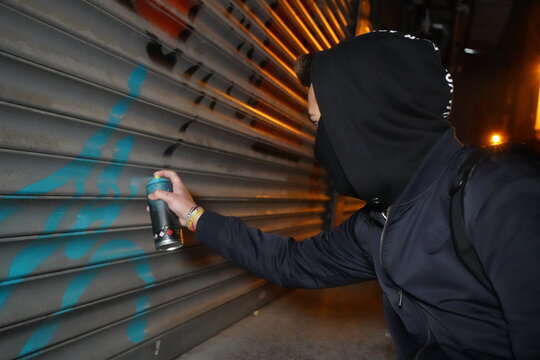 A young graffiti artist in a black hoodie looks at the wall, Street art concept, Vandal boy with hidden face makes painting graffiti on wall, Illegal Young man Spraying turquoise paint on a shutter.