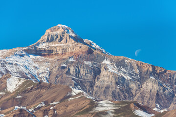 Mount Gestinkil close up, Dagestan, Russia. Mountain landscape with moon on clear blue sky. Alpine meadows and rocks covered with snow on a sunny day
