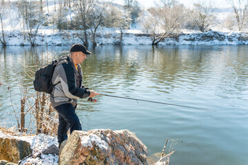 A fisherman with a fishing rod and a backpack catches fish on the bank of a snow-covered river in...