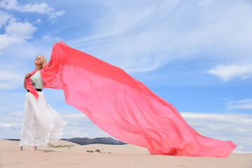Young woman standing in sand dunes with a wide and large coral pink color textile silk scarf around her on a windy day.