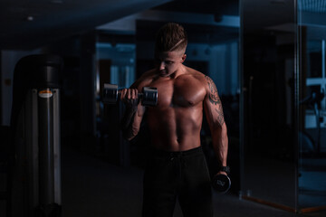 Fototapeta na wymiar Fitness man model with hairstyle and tattoo workout with dumbbell in gym in the dark. Sports lifestyle