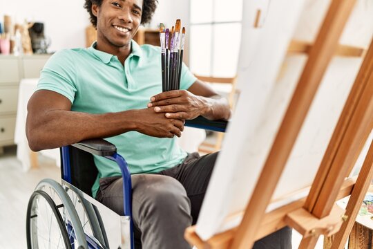 Young african american disabled artist man holding paintbrushes sitting on wheelchair at art studio.